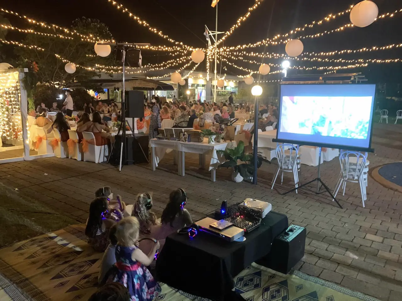 Children watching a movie on a big screen with silent headphones while the party continues behind them
